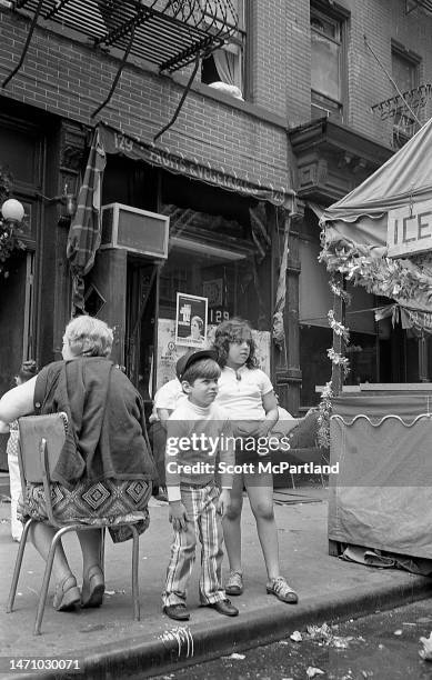 View of two children standing next to a senior adult seated in a chair on Mulberry Street , during the Feast Of San Gennaro Festival, in the Little...