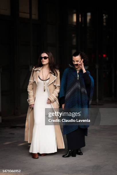 Evelina Maria Corco wears a cream lonng dress, tan trench coat with white details, Debi Mazar wears a blue and grey cape and black boots, outside...