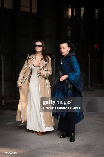 Evelina Maria Corco wears a cream lonng dress, tan trench coat with white details, Debi Mazar wears a blue and grey cape and black boots, outside...