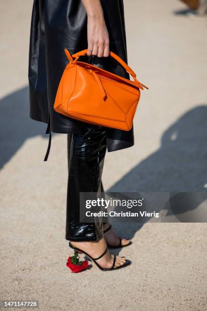 Xenia Adonts wears black sleeveless leather dress, orange Loewe bag, varnished pants, heeled sandals outside Loewe during the Paris Fashion Week -...