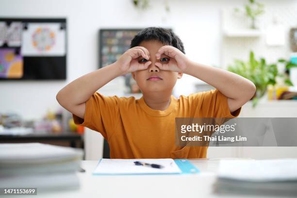 cheerful schoolboy  did fingers like binoculars while works on homework assignment - asian child with binoculars stockfoto's en -beelden