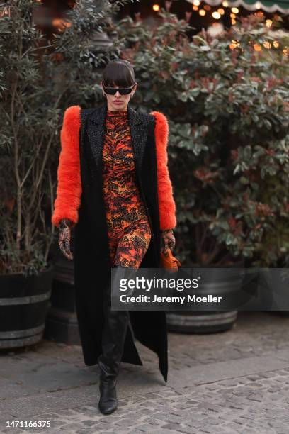 Katya Tolstova seen wearing black Pinko boots, Prada shades, an orange patterned dress by Marcell von Berlin, an orange mini bag, a black long coat...
