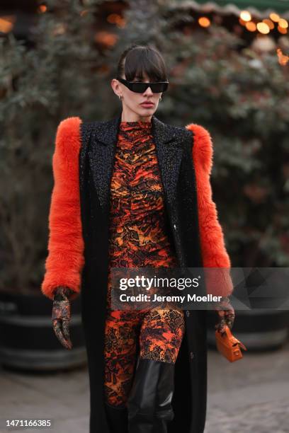 Katya Tolstova seen wearing black Pinko boots, Prada shades, an orange patterned dress by Marcell von Berlin, an orange mini bag, a black long coat...