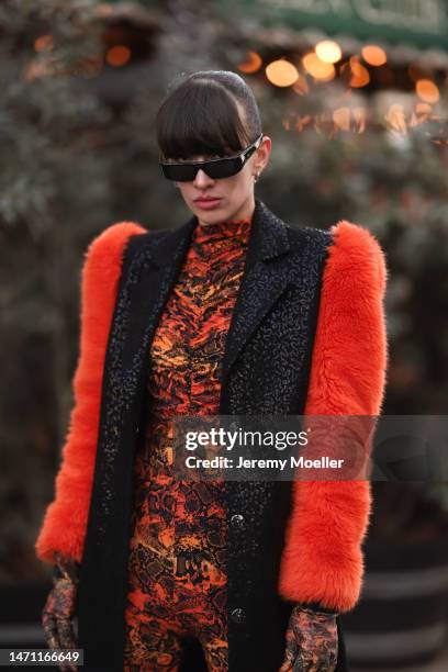 Katya Tolstova seen wearing black Pinko boots, Prada shades, an orange patterned dress by Marcell von Berlin, an orange mini bag, a black long coat...