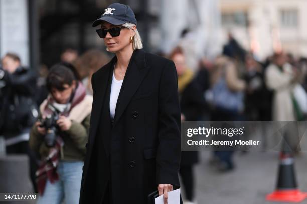 Fashion week guest seen wearing a cap by Celine, dark shades, a white shirt and a long black coat before the Chloe show on March 02, 2023 in Paris,...