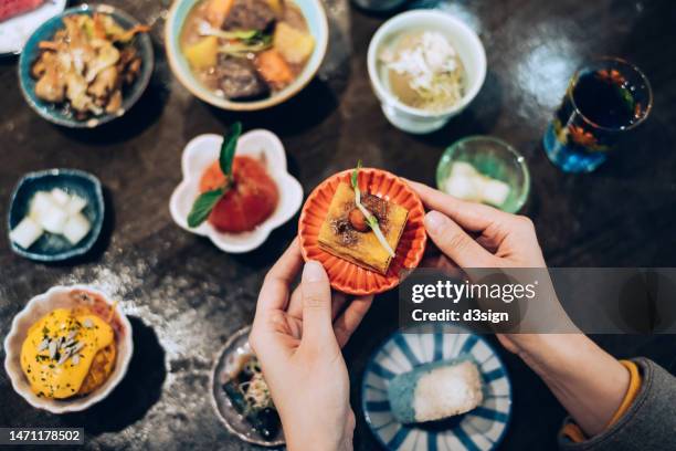 overhead view of a female hand holding a plate of japanese style tamagoyaki in hands, with various delicate side dishes on wooden dining table in background. authentic japanese cuisine. asian cuisine and food culture. people, food and lifestyle - serving dish stock pictures, royalty-free photos & images