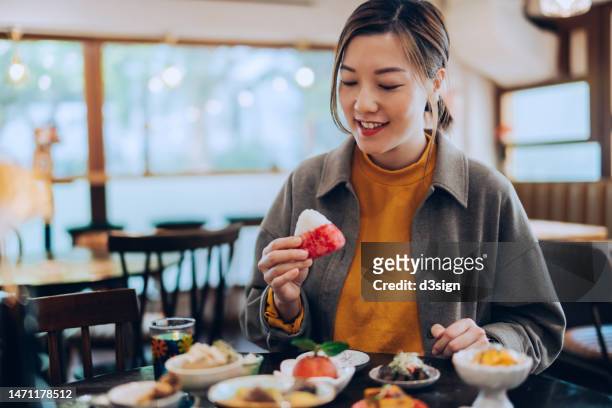 smiling young asian woman eating a mount fuji onigiri while enjoying delicate japanese cuisine, with scrumptious side dishes and green tea served on the table in restaurant. asian cuisine and food culture. people, food and lifestyle - luxury home dining table people lifestyle photography people stock pictures, royalty-free photos & images