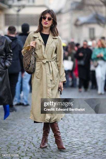 Jeanne Damas wears brown sunglasses, gold earrings, a black wavy cut-out pattern neck pullover, a beige long belted coat, a beige and black zebra...