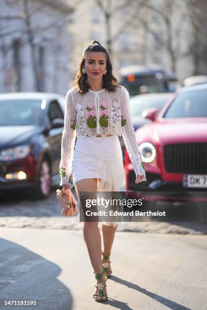 Alexandra Pereira wears silver and crystals large earrings, a white lace print pattern with embroidered pink and green flower pattern long sleeves...