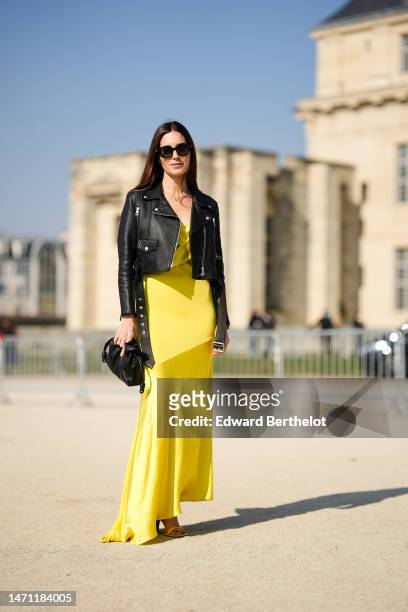 Gala Gonzalez wears black sunglasses, a black shiny leather zipper cropped jacket, a yellow V-neck / satin silk / long asymmetric dress, a black...