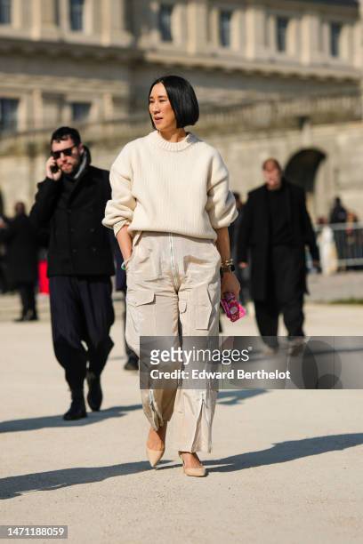 Eva Chen wears a white latte wool pullover, beige shiny satin cargo pants, gold bracelets, a gold watch, beige shiny leather pointed pumps heels...