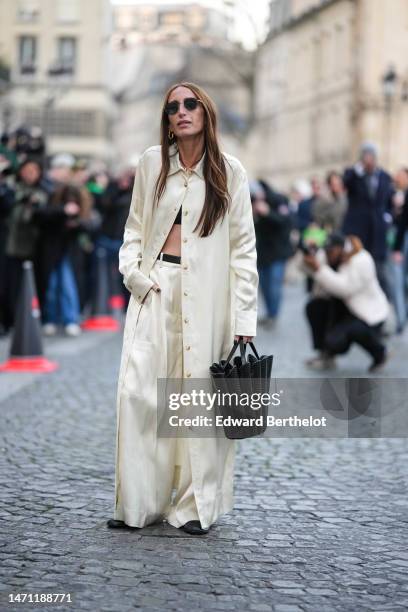 Chloe Harrouche wears black sunglasses, gold earrings, a black cropped top, a pale yellow / white latte long buttoned shirt, matching pale yellow /...