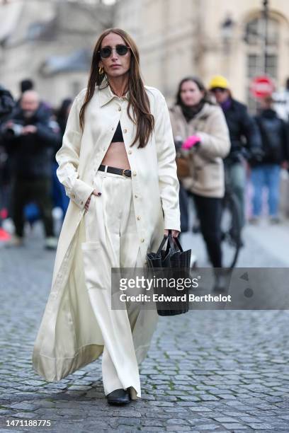 Chloe Harrouche wears black sunglasses, gold earrings, a black cropped top, a pale yellow / white latte long buttoned shirt, matching pale yellow /...