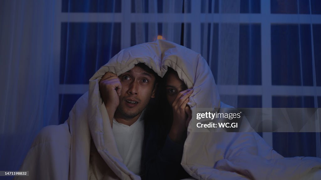 Asian couple watch horror movies on the bed with blanket to cover their heads