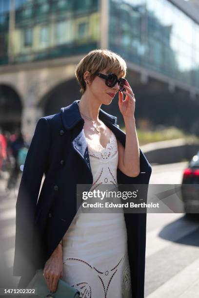 Rebecca Hall wears black large sunglasses, a navy blue wool long coat, a white V-neck lace print pattern midi dress with cut-out pattern, a pale...