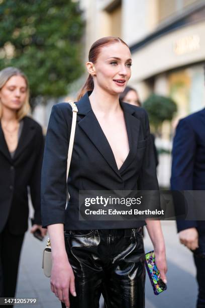 Sophie Turner wears gold earrings, a black blazer jacket, black shiny varnisged leather pants, a white shiny leather handbag, outside the LVMH...