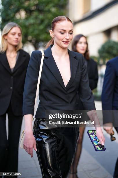 Sophie Turner wears gold earrings, a black blazer jacket, black shiny varnisged leather pants, a white shiny leather handbag, outside the LVMH...