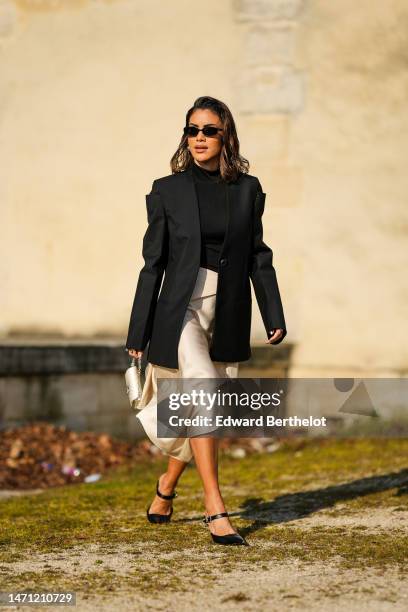 Camila Coelho wears black sunglasses, silver earrings, a black high neck / turtleneck t-shirt, a black oversized blazer jacket, a beige silk / satin...