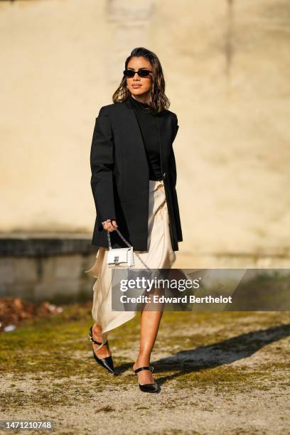 Camila Coelho wears black sunglasses, silver earrings, a black high neck / turtleneck t-shirt, a black oversized blazer jacket, a beige silk / satin...
