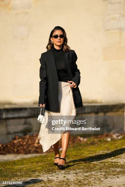 Camila Coelho wears black sunglasses, silver earrings, a black high neck / turtleneck t-shirt, a black oversized blazer jacket, a beige silk / satin...