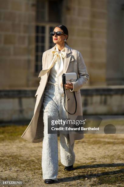 Julia Haghjoo wears black sunglasses, silver large pearl earrings, a white latte silk knot neck blouse, a beige long wool coat, a white embroidered...