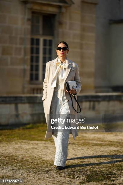 Julia Haghjoo wears black sunglasses, silver large pearl earrings, a white latte silk knot neck blouse, a beige long wool coat, a white embroidered...