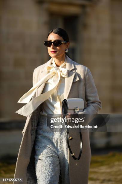 Julia Haghjoo wears black sunglasses, silver large pearl earrings, a white latte silk knot neck blouse, a beige long wool coat, a white embroidered...