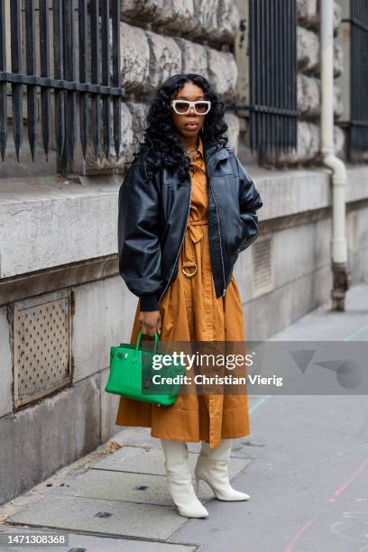 Guest wears black bomber jacket, brown dress, green Hermes bag, white boots, sunglasses outside Hermes during Paris Fashion Week - Womenswear Fall...