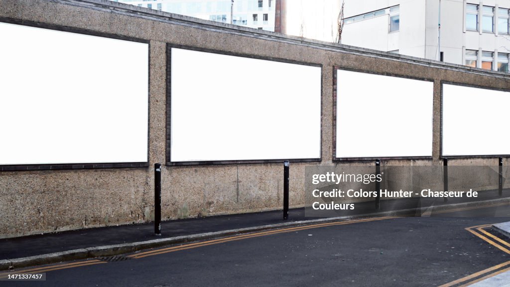 Four white and empty advertising panels on a weathered wall with sidewalk and street in London, England, United Kingdom