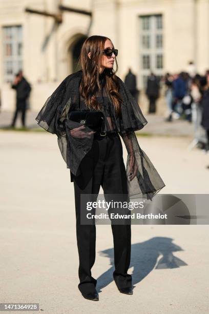 Chloe Harrouche wears black sunglasses from Tol, gold earrings, a black pleated / accordion tulle blouse with long ruffled sleeves , a black...