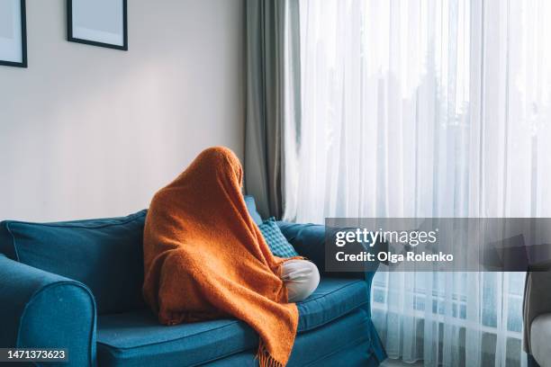 unrecognizable human covered by orange blanket, like a ghost on the blue sofa in living room - pms stock pictures, royalty-free photos & images