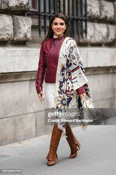 Nojoud Alrumaihi wears diamonds earrings, a white turtleneck t-shirt, a burgundy shiny leather asymmetric zipper jacket from Hermes, a white with...