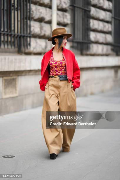 Guest wears a brown felt hat, gold and brown leather pendant earrings, a neon red wool zipper jacket, a purple with yellow and pink / orange print...