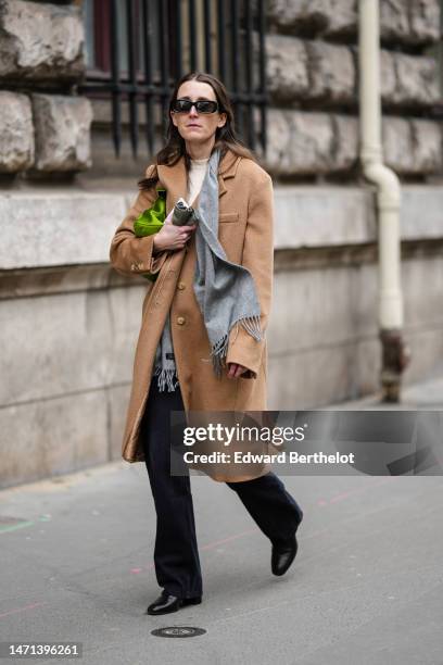 Guest wears black squared sunglasses, a beige t-shirt, a gray wool fringed scarf, a beige wool long coat, a green shiny satin puffy handbag, green...