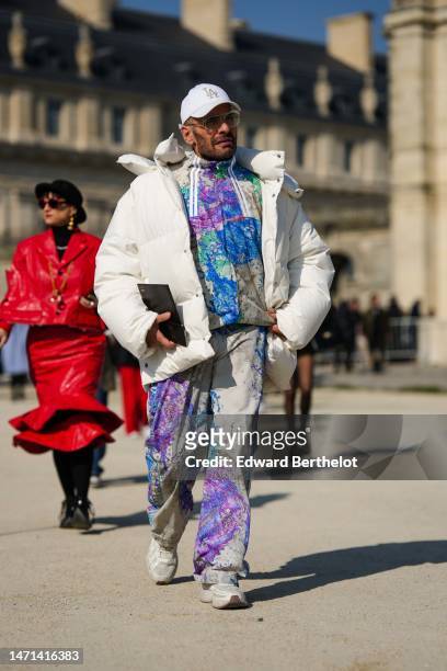Guest wears a white with gold embroidered LA cap , transparent glasses, a beige with purple / blue / green print pattern zipper jacket, matching...