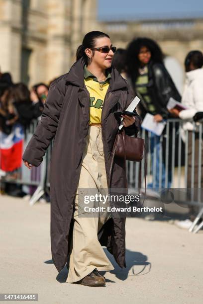 Guest wears brown sunglasses, a khaki polo shirt, a yellow with khaki print pattern t-shirt, a dark brown long puffer jacket, a beige long denim...