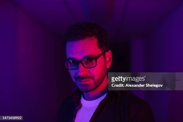 male portrait of man with neon lighting. red, blue, purple and magenta colours with dark background. person wear eyeglasses, dark jacket and white t-shirt indoors. futuristic lifestyle concept multicolour light - red light portrait stock pictures, royalty-free photos & images