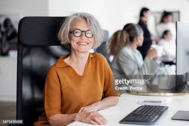 portrait of a cheerful senior businesswoman sitting at office desk - 60 64 years stock pictures, royalty-free photos & images