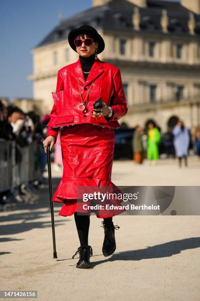 Guest wears a black felt wool hat, red sunglasses, a black wool turtleneck pullover, a gold necklace, a red shiny leather ripped jacket, a red shiny...