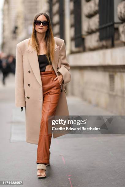 Charlotte Groeneveld wears black large sunglasses, a beige wool long coat, black cut-out waist body, camel shiny leather large pants, beige puffy...
