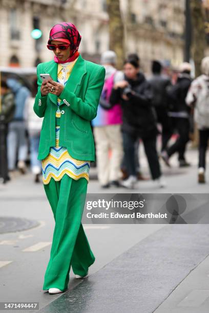 Guest wears a red and black print pattern silk scarf as a headband, a yellow / blue / white embroidered wavy pattern long t-shirt, a green asymmetric...