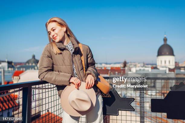 Fashionable young woman enjoying landscape from balcony of European city holding hat. Spring fashion