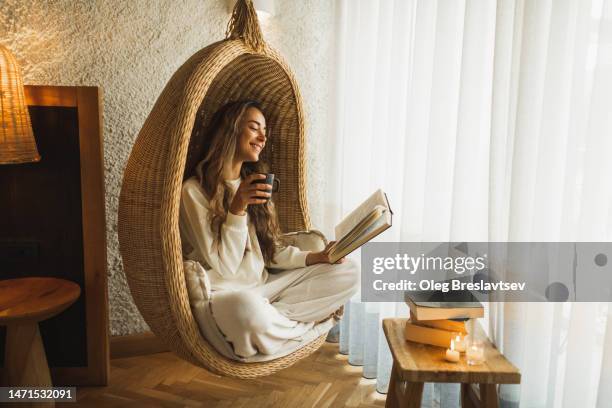 woman relaxing at home in hanging ball chair, reading book and drinking hot herbal tea - bubble chair stock-fotos und bilder