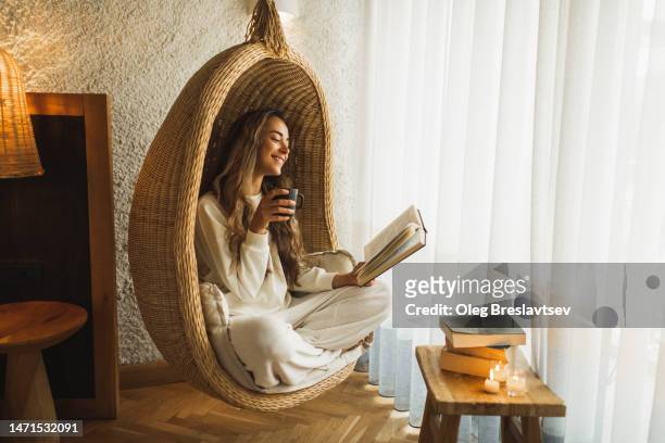 woman relaxing at home in hanging ball chair, reading book and drinking hot herbal tea - egg chair stockfoto's en -beelden