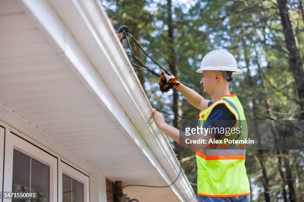 junge erwachsene arbeiter reinigen blätter von einer dachrinne auf einer hohen leiter. konzentrieren sie sich auf den vordergrund mit defokussiertem hintergrund. - leaf on roof stock-fotos und bilder