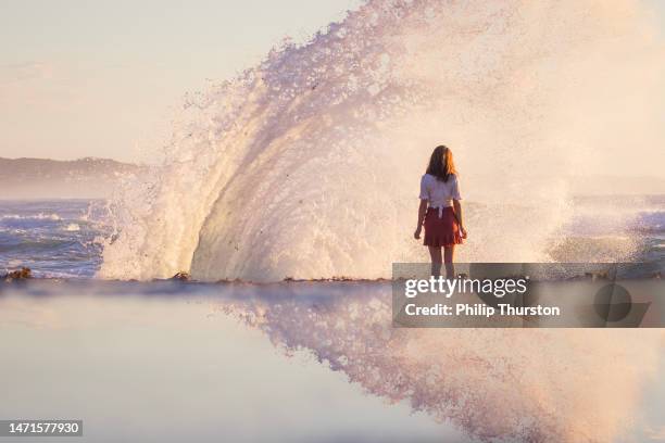 young woman in dress standing by the ocean as waves crash and spray in golden light around her - gold meets golden stock pictures, royalty-free photos & images