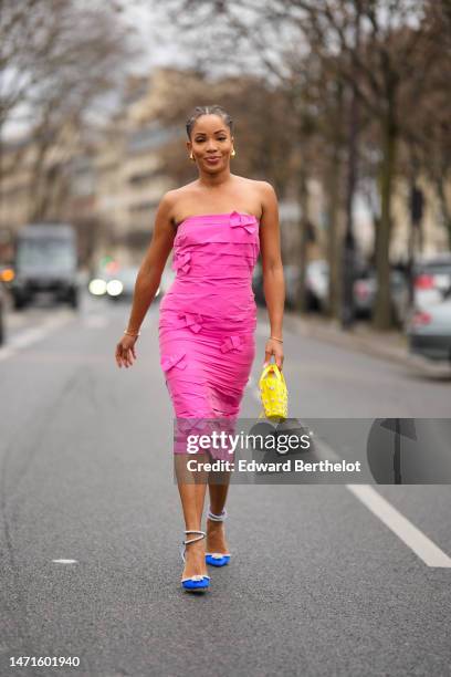 Ellie Delphine wears gold earrings, a neon pink shoulder-off / belted midi tube dress, a yellow shiny leather with embroidered crystals flower...