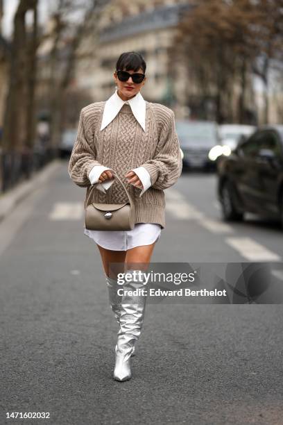 Gili Biegun wears black sunglasses, gold and white pearls pendant earrings, a white shirt with large collar short shirt dress, a brown braided wool...