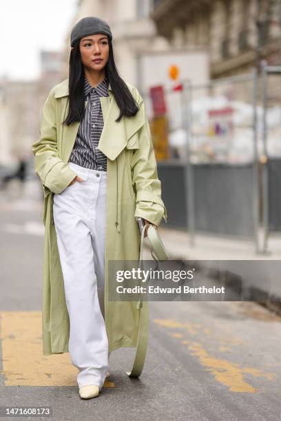 Yoyo Cao wears a dark gray felt wool beanie, a dark gray and white striped print pattern shirt, a pale green shiny leather long belted trench coat,...