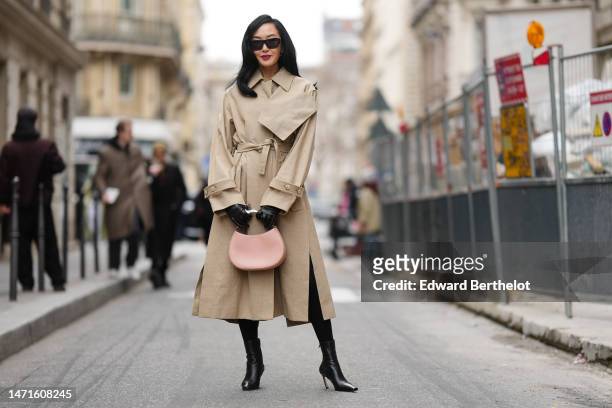 Chriselle Lim wears black sunglasses, a beige linen long belted trench coat, black shiny leather gloves, a pale beige pink shiny leather with silver...